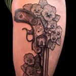 Фото тату: черно-белый рисунок револьвера, окруженного цветами и листьями на руке. Детализированная татуировка с использованием теней.