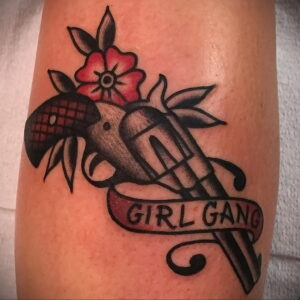 Фото тату револьвера с розой и надписью 'Girl Gang' на предплечье. Цветной рисунок татуировки в стиле олд скул с четкими контурами и яркими цветами.