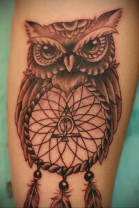 Фото тату совы в стиле blackwork, с детально проработанным ловцом снов и перьями, расположенной на руке.
