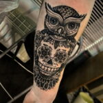 Фото татуировки на руке в стиле blackwork с изображением совы, переходящей в сахарный череп. Четкие линии и контрастный дизайн.