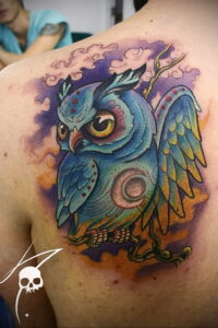 Фото акварельной татуировки совы на плече, выполненной в нежных пастельных тонах с использованием техники размытия. Красивый рисунок татуировки.