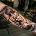 Фото тату: реалистичное изображение двух сцепленных кулаков, выполненное в черно-серых тонах на предплечье мужчины. Детализированный рисунок татуировки.