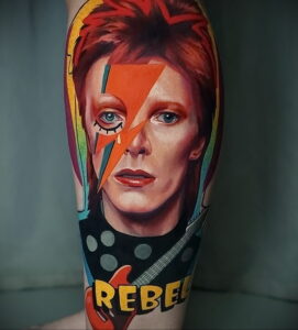 Фото тату: реалистичный портрет Дэвида Боуи в его знаменитом образе Ziggy Stardust на руке, с изображением гитары и надписи 'Rebel'.