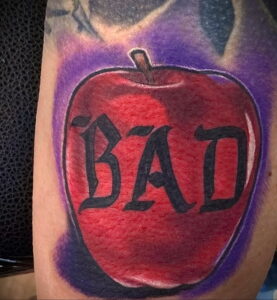 Фотография татуировки в виде красного яблока с надписью 'BAD' на руке. Рисунок выполнен в ярких цветах с использованием теней и бликов.