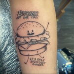 Фото тату: простой черно-белый бургер с надписью 'Burger of the Day' и фразой 'It's only skin deep', расположенный на руке.