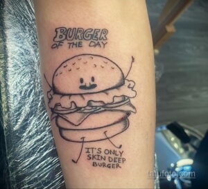 Фото тату: простой черно-белый бургер с надписью 'Burger of the Day' и фразой 'It's only skin deep', расположенный на руке.