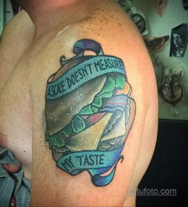 Фотография татуировки бургера с надписью 'A scale doesn't measure my taste'. Рисунок выполнен в реалистичном стиле с использованием ярких цветов и теней.