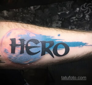 Фото татуировки на руке с надписью "HERO", выполненной в сине-голубых тонах с эффектом акварели и космического фона. Оригинальный и вдохновляющий рисунок татуировки.
