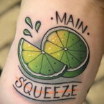 Фото тату на руке: половинка лайма с надписью 'Main Squeeze'. Яркие цвета, четкие контуры, традиционный стиль.