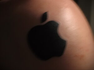 Фото тату: стилизованное изображение логотипа Apple на плече, выполненное в черном цвете. Современный и минималистичный рисунок татуировки.