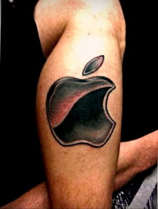 Фото тату на бицепсе: стилизованное изображение логотипа компании Apple, выполненное в черно-серых тонах.