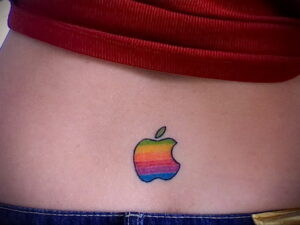 Небольшая татуировка на пояснице: стилизованное изображение логотипа Apple в виде надкушенного яблока, выполненное в цветах радуги. Яркий и минималистичный рисунок татуировки.