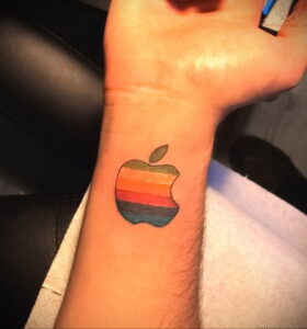 Небольшая татуировка на запястье: стилизованное изображение логотипа Apple в виде надкушенного яблока, выполненное в ярких цветах радуги. Минималистичный рисунок татуировки.