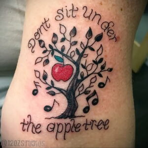 Фото татуировки яблони с надписью 'Don't sit under the apple tree' и музыкальными нотами. Черно-белый рисунок татуировки на лодыжке.