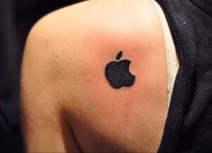 Фото тату на плече: маленький черный логотип Apple. Минималистичный и лаконичный дизайн.