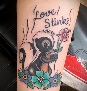Фото тату скунса, нюхающего цветок, с надписью 'Love Stinks', выполненное в ярком и красочном стиле.