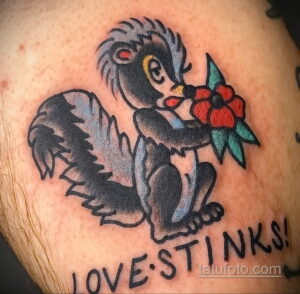 Фото тату скунса с розой и надписью "LOVE STINKS!" на руке. Черно-серый рисунок в стиле олдскул с яркой надписью.