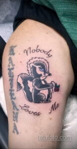 Фото тату скунса с грустной надписью 'Nobody Loves Me' на плече. Рисунок выполнен в черно-белой гамме с добавлением красного цвета.