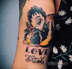 Фото тату с изображением енота в стиле олд скул и надписью "Love Stinks" на руке. Яркие цвета, четкие контуры и провокационный посыл.