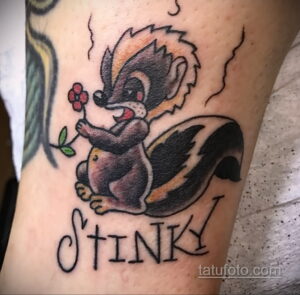 Фото тату енота с розочкой и надписью 'Stinky' на руке. Стиль олд скул, яркие цвета, милый и ироничный дизайн.