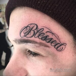 Фото тату надписи 'Blessed' на лбу, выполненное черными линиями в каллиграфическом стиле. Татуировка расположена над бровью.