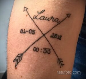 Фото тату на руке: минималистичная надпись "Laura" с датой "01-05-2018" и временем "00:33", оформленная в виде стрелы. Черно-белый рисунок татуировки.