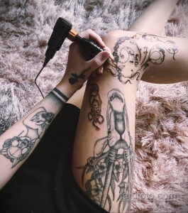 Фото тату: Мастер делает татуировку в стиле Blackwork на бедре клиентки. Видны татуировки на руке мастера и процессе нанесения рисунка.