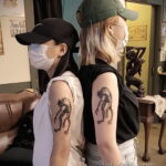 Фото двух людей с одинаковыми татуировками странных существ на плече. Изображения выполнены в черном цвете, с четкими контурами и минимальной детализацией.
