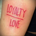 Фото минималистичного тату с надписью "LOYALTY" и "LOVE", перечеркнутыми красной линией, на запястье.