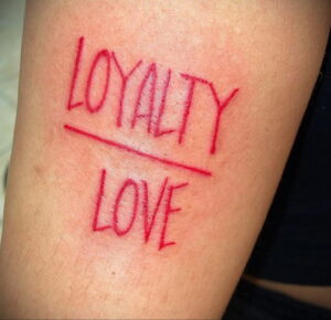 Фото минималистичного тату с надписью "LOYALTY" и "LOVE", перечеркнутыми красной линией, на запястье.