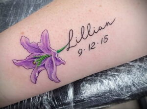 Фото тату на предплечье, изображающее лилию и надпись "Lillian 9.12.15". Цветочная татуировка с датой, символизирующая память.