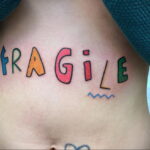 Фото тату на боку, изображающее надпись "FRAGILE" разноцветными буквами. Необычный и привлекающий внимание дизайн.