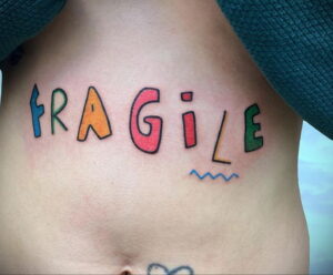 Фото тату на боку, изображающее надпись "FRAGILE" разноцветными буквами. Необычный и привлекающий внимание дизайн.