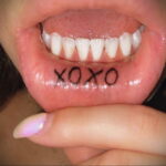 Фото тату с надписью 'XOXO' внутри рта, на нижней губе. Миниатюрный рисунок, выполненный черными чернилами, символизирует любовь и объятия.