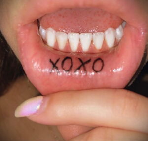 Фото тату с надписью 'XOXO' внутри рта, на нижней губе. Миниатюрный рисунок, выполненный черными чернилами, символизирует любовь и объятия.