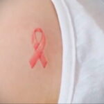 Фото тату: небольшая розовая лента, символизирующая поддержку, на плече. Минималистичный дизайн, акварельная техника.