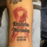Фото тату на руке: красная лента с именем 'Rafaela Morales' и датами, выполненное в стиле акварели.