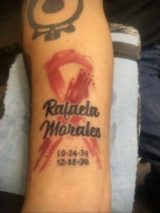 Фото тату на руке: красная лента с именем 'Rafaela Morales' и датами, выполненное в стиле акварели.