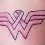 Фото тату: на руке изображен символ Wonder Woman, в центре которого расположена розовая лента. Символ силы и борьбы.