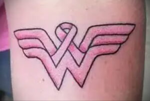 Фото тату: на руке изображен символ Wonder Woman, в центре которого расположена розовая лента. Символ силы и борьбы.