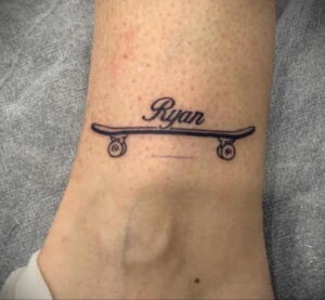 Фото тату на лодыжке с изображением скейтборда и надписью 'Ryan'. Стиль - минимализм, черные линии, простой дизайн.