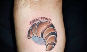 Фото тату круассана с надписью "Cornetto!!!", выполненного в ярком стиле с использованием теплых коричневых и красных оттенков.