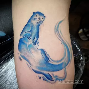 Фото акварельной татуировки с изображением синей выдры, плывущей в воде. Тату выполнено в мягких, размытых тонах, создающих эффект движения.