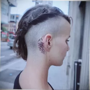 Фото нежной татуировки с изображением ветки сирени за ухом. Тонкие линии, минималистичный дизайн, изящное расположение.