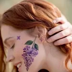 Фото тату: реалистичная татуировка ветки сирени с распустившимися цветами, расположенная на щеке девушки с рыжими волосами.