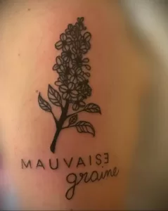 Фото тату с изображением ветки сирени и надписью "Mauvaise Graine" на французском языке. Черно-белый рисунок с каллиграфическим шрифтом.