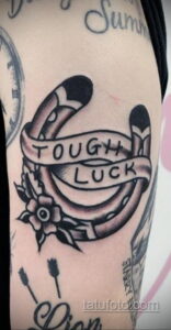 Фото тату подковы с надписью "TOUGH LUCK" и цветком в стиле олд скул. Яркие цвета, четкие контуры, традиционный дизайн. Расположена на плече.