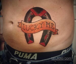 Фото татуировки подковы с надписью 'Lucky Me', выполненной в традиционном стиле с использованием красного и черного цветов на животе.
