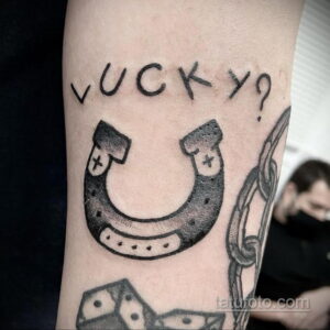 Фото тату подковы с надписью 'Lucky?' на руке. Черно-белый рисунок в минималистичном стиле с добавлением цепи.