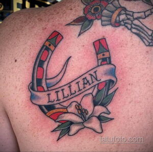 Фото тату подковы с надписью 'Lillian' и цветком лилии на плече. Яркий рисунок в стиле олд скул с использованием красного, синего и зеленого цветов.